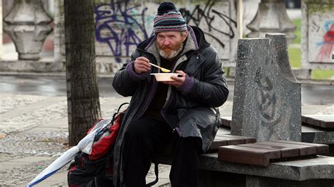 Bezdomovec sedí na lavičke v parku s taškami, v pozadí prechádzajú ľudia